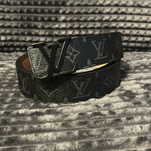 Louis Vuitton svart skinnbälte - Snyggt svart bälte från Louis Vuitton med klassiskt monogrammönster och stor LV-logga i metall på spännet. Bältet är gjort i exklusivt skinn och har en stilren, mörk look som lyfter vilken outfit som helst.