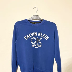 Blå Calvin Klein tröja med tryck - Snygg blå tröja från Calvin Klein Jeans med stort vitt tryck på bröstet där det står 'CALVIN KLEIN CK NEW YORK'. Tröjan har rund hals, långa ärmar och är gjord i mjuk bomull. Perfekt för dig som gillar klassisk streetstyle med en modern touch.