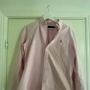 Rosa och vit randig skjorta Polo Ralph Lauren - Snygg rosa och vit randig skjorta från Polo Ralph Lauren med klassisk krage och knappar framtill. Långärmad modell med broderad logga på bröstet. Perfekt för dig som gillar stilrena och tidlösa plagg.ny pris 1600kr säljer endast för 499kr