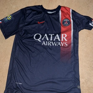 PSG Messi 30 matchtröja Nike - Snygg Paris Saint-Germain fotbollströja med Messi och nummer 30 på ryggen. Tröjan är marinblå med röd och vit rand på framsidan, PSG-logga på bröstet och Qatar Airways som sponsor. Tillverkad i lätt, ventilerande material med korta ärmar och rund hals.