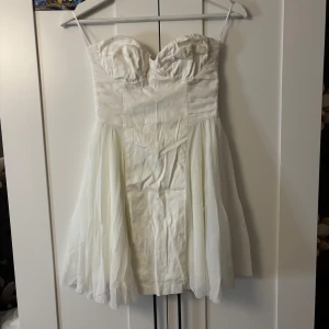 Studentklänning  - Supersöt vit axelbandslös klänning med hjärtformad byst och voluminös kjol. Klänningen har dragkedja bak och är tillverkad i bomull med polyesterfoder och meshdetaljer. Perfekt för dig som vill ha en romantisk och fräsch look. Passar jättebra till studenten, och skulle säga att bysten är B-C kupa. Behöver strykas innan användning.