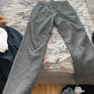 Grå mjukisbyxor från Champion - Gråa mjukisbyxor från Champion med elastisk midja och avslappnad passform. Byxorna har ett litet Champion-logo broderat på vänster lår. Perfekta för chill eller träning, mjukt material och klassisk look.