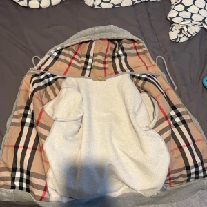 Grå kofta från Burberry, S - Säljer en grå teddyjacka från Burberry med klassiskt rutigt foder i beige, svart och rött. Jackan har huva, dragkedja och broderad logga på bröstet. Insidan är mjuk och fluffig, perfekt för kyliga dagar. Riktigt snygg och bekväm jacka med exklusiv känsla. Pris Ej hugget i sten så det är bara att komma med förslag!