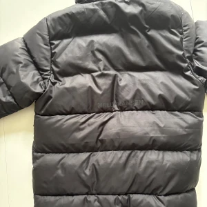 Svart pufferjacka Helly Hansen 152/12 - Svart pufferjacka från Helly Hansen med hög krage och två sidofickor. Jackan har quiltad design och dragkedja framtill. Perfekt för kalla dagar och riktigt skön att bära. Klassisk look med diskret HH-logga på bröstet. Den passar alltifrån 145-155. Priset är inte hugget i sten