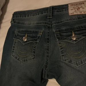 True Religion Becca Bootcut Jeans - Snygga blå jeans från True Religion, modell Becca Mid Rise Bootcut. Klassiska bakfickor med lock och stora silverfärgade knappar samt gul kontrastsöm. Jeansen har bootcut-passform och är tillverkade i slitstarkt denimtyg. Helt nya prislappen är kvar. De va i storlek 28 men sydde om de till storlek 23-25-25 och jag har sytt de till low Waits från mid 
