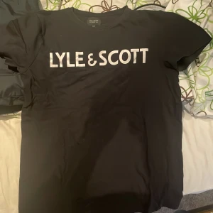 Svart Lyle & Scott t-shirt med tryck - Svart t-shirt från Lyle & Scott Junior med stort vitt logotryck på bröstet. Klassisk rund hals och korta ärmar. T-shirten är gjord i mjuk bomull och har en clean, enkel look som passar perfekt till jeans eller shorts.