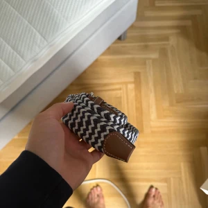 Randigt flätat bälte med skinn - Säljer ett snyggt flätat bälte med randigt mönster i svart och vitt. Bältet har detaljer i brunt skinn vid ändarna och är flexibelt i formen. Perfekt för att ge outfiten lite extra edge med sin coola design och kontrasterande färger.