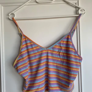 Randig ribbad linnetopp i pastell - Superfin ribbad linnetopp med smala axelband och färgglada ränder i blått, orange, rosa och gult. Toppen har en v-ringad hals och croppad passform, perfekt för varma dagar och festivalvibbar.