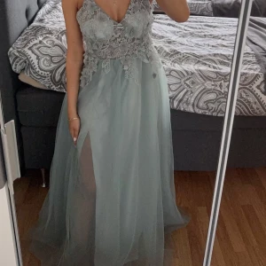 Bal/festklänning  - Säljer en drömmig ljusblå/grön långklänning med tunna axelband, v-ringning och vackra spetsapplikationer upptill. Kjolen är i flera lager tyll med en hög slits och öppen rygg med snörning. Perfekt för fest eller bal. 