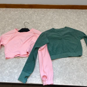 Två croppade träningströjor i rosa och grönt - Säljer två croppade långärmade träningströjor. Ena är helrosa och den andra är grön med rosa ärm. Båda har tumhål i ärmsluten och är gjorda i stretchigt material, perfekta för gymmet eller löpning.