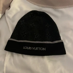 Svart Louis Vuitton mössa med logga - Snygg svart mössa från Louis Vuitton med ikoniskt monogrammönster och broderad logga framtill. Mössan har en grå kant och är tillverkad i mjukt material som känns lyxigt. Perfekt accessoar för dig som gillar exklusiv streetstyle.