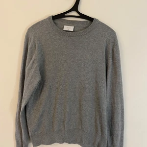 Grå stickad tröja från Grunt XL - En enkel och stilren grå stickad tröja från Grunt i storlek XL i barnstorlek. Den passar för personer som är mellan 165-172 cm långa ungefär. Tröjan har rund halsringning, ribbade muddar och är långärmad. Perfekt för lager-på-lager och passar till många olika outfits.