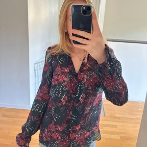 Blommig svart och röd blus - Säljer en snygg blus med svart bas och röda blommiga mönster. Blusen har v-ringning, långa ärmar med resår vid handlederna och är gjord i ett lätt och luftigt material. Perfekt att styla med jeans för en cool look. 