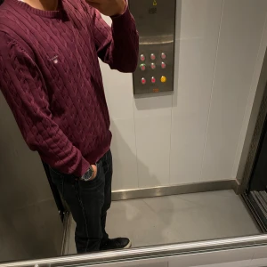 Vinröd kabelstickad tröja från Gant - Vinröd kabelstickad tröja från Gant med rund halsringning och diskret broderad logga på bröstet. Tröjan har lång ärm och ribbade muddar vid ärmslut och nederkant. Perfekt att bära över skjorta för en klassisk look.