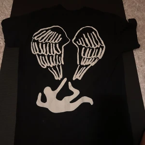 Svart Lil Nas X t-shirt med vingar - Svart t-shirt från Lil Nas X med coolt tryck på ryggen: vita änglavingar och en siluett. Framtill finns en liten vit logga och vinge. Klassisk passform och rund hals. Perfekt för dig som gillar statement-plagg och streetwear.