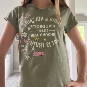 Grön t-shirt Zadig & Voltaire med tryck - Cool grön t-shirt från Zadig & Voltaire med vit och rosa tryck framtill, blommor, stjärnor, peace-symboler och text. Rund hals med rå kant och två knappar. Skön passform och tillverkad i 100% bomull. Perfekt för dig som gillar statement-plagg.