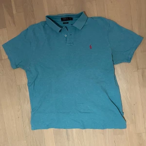 Blå Polo Ralph Lauren piké - Snygg blå piké från Polo Ralph Lauren med klassisk pikékrage och två knappar framtill. Den har korta ärmar och en röd broderad logga på bröstet. Materialet är mjuk bomull som känns skönt mot huden. Perfekt för dig som gillar stilren och enkel design.