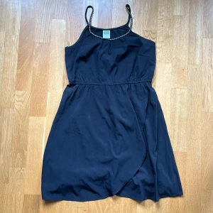 Svart omlottklänning Vero Moda - Snygg svart kortklänning från Vero Moda med omlottkjol och tunna axelband dekorerade med pärlor. Klänningen har en lätt och luftig känsla, perfekt för varma dagar. Stilren design som ger en cool och avslappnad vibe.