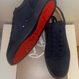 Svarta sneakers från Christian Louboutin - Snygga svarta sneakers i mocka från Christian Louboutin med ikonisk röd sula. Skorna har rund tå, svarta snören och diskret broderad logga på hälen. Perfekt för dig som vill sticka ut med exklusiva detaljer och lyxig känsla.