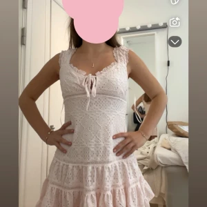 Rosa spetsklänning med volanger - Supersöt ljusrosa kortklänning med spetsdetaljer och volanger. Klänningen har snörning framtill vid urringningen och är ärmlös med breda axelband. Perfekt för sommarens alla tillfällen, med romantisk och feminin vibe.