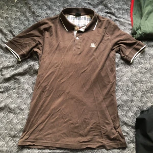 Brun Burberry pikétröja med logga - Snygg brun pikétröja från Burberry med klassisk krage, vita detaljer på krage och ärmslut samt broderad logga på bröstet. Insidan av kragen har det ikoniska rutmönstret. Perfekt för en stilren och avslappnad look.