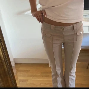 Beige lågmidjade jeans  - Säljer ett par snygga beige byxor med låg midja. Byxorna har markerade sömmar framtill och coola fickor med lock och knappdetaljer. Aldrig använda. Helt helt nya men har tagit bort lappen. 