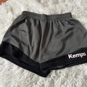 Grå sportshorts från Kempa - Snygga grå sportshorts från Kempa med svart kant nertill och elastisk midja. Perfekta för träning eller sport, tillverkade i lätt polyester som andas. Loggan syns tydligt på benet och modellen är loose fit för skön rörelsefrihet.