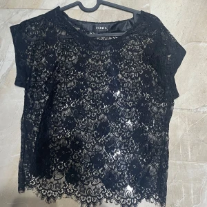 Svart spetsblus från Club L - Säljer en svart blus från Club L med vacker spets och korta ärmar. Blusen har rund halsringning och ett genombrutet spetsmönster som ger en elegant look. Perfekt att styla med jeans eller kjol för en trendig outfit.