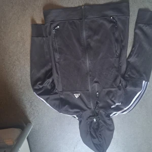Svart Adidas hoodie med dragkedja - Svart hoodie från Adidas med hel dragkedja, huva och två fickor med dragkedja framtill. Klassiska vita ränder längs ärmarna och Adidas-logga på bröstet. Perfekt för en sportig look eller chill vardagsstil.