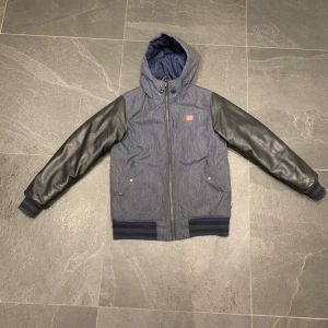 Blå bomberjacka med svarta ärmar - Snygg blå vinterjacka  med svarta skinnärmar och huva. Jackan har dragkedja framtill, ribbade muddar med blå och svarta ränder samt två fickor med tryckknapp. Perfekt för dig som vill ha en streetig look. Den är x small och passar dig som är lite kortare