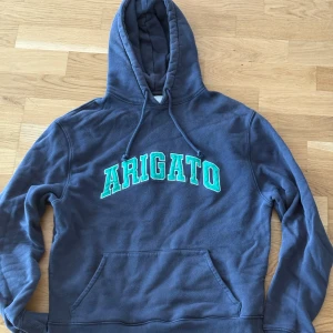 Axel Arigato hoodie  - Snygg mörkblå hoodie från Axel Arigato med stor grön och vit ARIGATO-text på bröstet. Klassisk känguruficka, huva med snörning och mjukt bomullsmaterial. Perfekt för en chill och trendig look. Storlek XS, hör gärna av er vid frågor :)