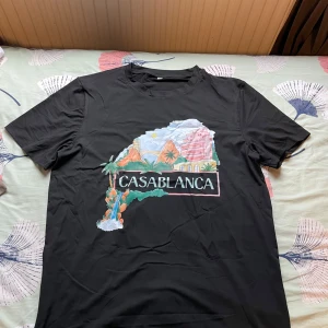 Svart Casablanca t-shirt med tryck - Säljer en svart t-shirt från Casablanca med färgglatt tryck framtill som visar ett landskap med palmer, berg och byggnader. T-shirten har rund hals och korta ärmar. Perfekt för dig som gillar statement-plagg och snygga prints.