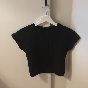 Svart croppad topp från Zara - En lite finare topp som man kan ha nu i vinter och höst, men också på sommaren en en vit kort kjol eller shorts!! Svart topp från Zara i storlek S med croppad passform och korta ärmar. Materialet är strukturerat och ger en cool textur. Rund halsringning och loose fit gör den enkel att styla med jeans eller kjol. Perfekt för dig som gillar minimalistisk stil.