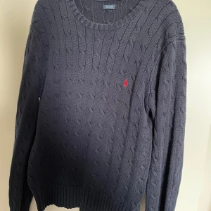 Mörkblå kabelstickad tröja Polo Ralph Lauren - Kabelstickad mörkblå tröja från Polo Ralph Lauren i 100% bomull. Klassisk rund halsringning och lång ärm, med den ikoniska röda broderade loggan på bröstet. Perfekt för dig som gillar stilren och tidlös design.