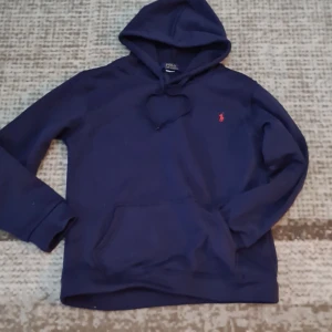 Marinblå hoodie från Polo Ralph Lauren - Snygg marinblå hoodie från Polo Ralph Lauren med klassisk röd broderad logga på bröstet. Tröjan har huva med snörning, känguruficka och är tillverkad i mjuk bomull. Perfekt för en chill och stilren look.