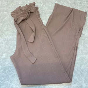 Beige ribbade byxor med knytband - Snygga ribbade byxor i beige med hög midja och volangkant upptill. Byxorna har ett matchande knytband i midjan och är i en mjuk, stretchig kvalitet. Perfekta för en chill och trendig look.