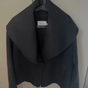 Kort jacka  Stylein med stor krage - Säljer en svart kappa från Stylein i storlek S. Jackan har en oversized, rundad krage som ger en cool och modern look. Tillverkad i ett tjockare ullmaterial, perfekt för kyligare dagar. Enkel och stilren design som lyfter vilken outfit som helst. Sparsamt använd köpt för 5000 kr