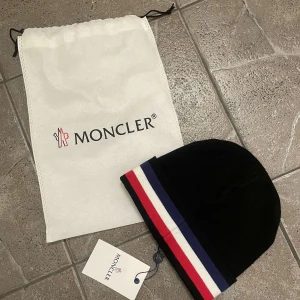 Svart Moncler mössa med ränder - Svart Moncler mössa i mjuk ull med klassiska Moncler-loggan framtill. Mössan har breda ränder i rött, vitt och blått längs kanten för en sportig och stilren look. Perfekt accessoar för att hålla värmen och lyfta din outfit.