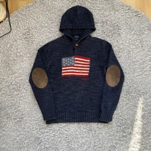 Mörkblå stickad hoodie Polo Ralph Lauren - Snygg mörkblå stickad hoodie från Polo Ralph Lauren med amerikansk flagga framtill, RL67-broderi och bruna mockadetaljer på armbågarna. Tröjan har huva, lång ärm och en träknapp vid halsen. Perfekt för dig som gillar klassisk och sportig stil. Storlek L i barn (sitter som S)