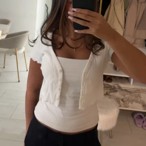 Vit topp  - Säljer en vit croppad topp från H&M Divided i storlek XS. Toppen har korta ärmar, knappar framtill och en söt, lätt ribbad struktur. Perfekt för dig som gillar en enkel men trendig look med lite edge.