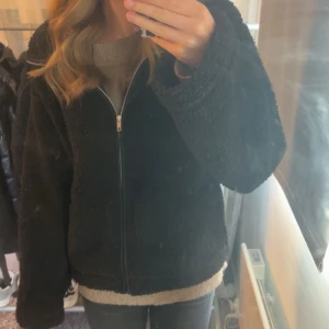 Svart teddyjacka med dragkedja - Mysig svart teddyjacka med dragkedja framtill. Jackan har en avslappnad passform och är perfekt för kyliga dagar. Den fluffiga ytan ger en cool och trendig look som passar till jeans eller leggings.