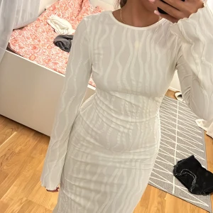 Vit långklänning med öppen rygg och rosett - Supersnygg vit långklänning med vågigt mönster, långärmad och figurnära passform. Klänningen har öppen rygg med en stor rosett som detalj och är tillverkad i ett lätt, stretchigt material. Perfekt för dig som vill sticka ut med stilren vibe.