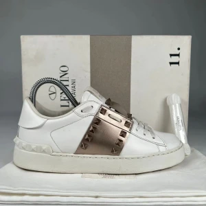 Valentino Rockstud sneakers vit/guld - Snygga Valentino Rockstud sneakers i vitt skinn med metallicguld band och ikoniska nitar. Klassisk rund tå, platt sula och snörning. Perfekt för dig som vill sticka ut med exklusiva detaljer och lyxig känsla.