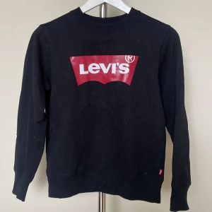 Svart Levi's sweatshirt med röd logga - Svart sweatshirt från Levi's med den klassiska röda loggan framtill. Tröjan har rund halsringning, långa ärmar och ribbade muddar vid ärmslut och nederkant. Perfekt för en chill och stilren look.