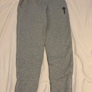 Grå trapstar byxa med en svart T loga på låret - Snygga grå mjukisbyxor med en svartvit T-logga på låret. Byxorna har dragkedja vid bensluten och detaljer i perforerat tyg på baksidan av benen. Perfekta för chill eller träning, med skön passform och elastisk midja.