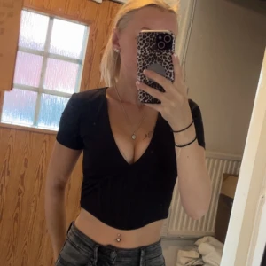 Svart croppad v-ringad topp - Säljer en svart croppad topp med djup v-ringning och korta ärmar. Toppen har en figurnära passform och är korsett liknande framtill som ger en snygg siluett. Perfekt att styla med höga jeans för en trendig look.