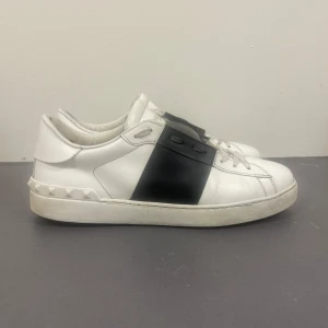 Valentino open - Snygga vita sneakers från Valentino Garavani med svart band över snörningen och ikoniska nitar på hälen. Skorna är i skinn och har en klassisk rund tå och platt sula. Perfekta för dig som vill ha en stilren men ändå unik look.
