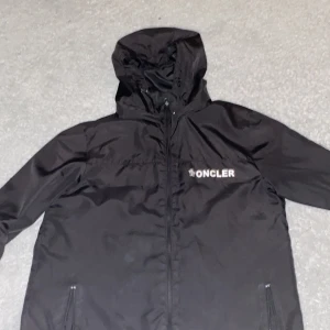 Svart vindjacka från Moncler - Svart vindjacka från Moncler med huva och dragkedja framtill. Jackan har Moncler-logga på bröstet och ärmen, samt två fickor med dragkedja. Snygg och stilren design i lätt material, perfekt för blåsiga dagar.