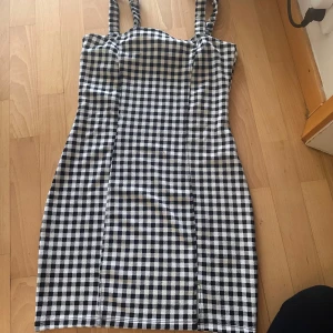 Svartvit rutig kort klänning - Säljer en svartvit rutig klänning med axelband och figurnära passform. Klänningen är kort och har ett klassiskt rutmönster som ger en cool och trendig vibe. Perfekt för dig som gillar stilrena och tidlösa plagg med lite edge.
