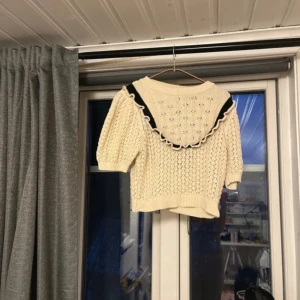 Beige stickad topp med volang från Zara - Supersöt beige stickad topp från Zara med korta ärmar och öppen rygg. Volangkant med svart detalj runt den öppna ryggen och ett diskret hålmönster över hela tröjan. Perfekt för lager-på-lager och snygg till både jeans och kjol.